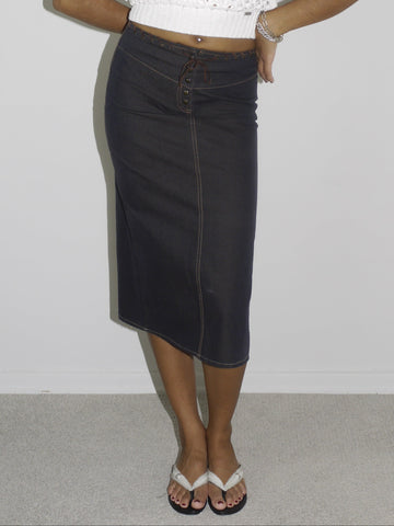 Model wearing a cropped white open-knit mesh top layered over a black bra, paired with a high-waisted dark denim midi skirt featuring contrast stitching and a wrap-effect detail. Styled with flip-flops and silver jewelry for a modern twist on early-2000s streetwear. Shot against a plain white background.