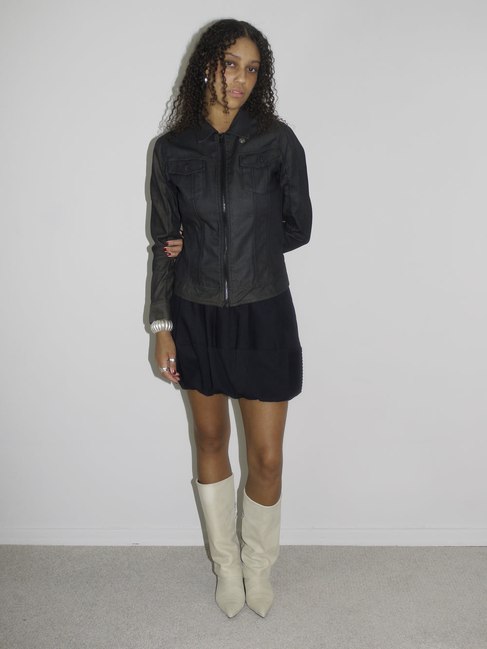Model wearing a black fitted denim jacket with front zip and chest pockets, styled over a voluminous black mini skirt. Finished with cream-colored pointed cowboy boots, blending tough outerwear with playful western influence. Shot against a plain white wall.