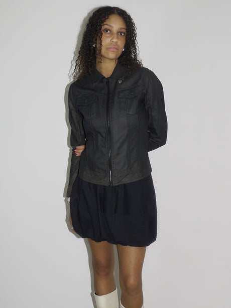 Model wearing a black fitted denim jacket with front zip and chest pockets, styled over a voluminous black mini skirt. Finished with cream-colored pointed cowboy boots, blending tough outerwear with playful western influence. Shot against a plain white wall.