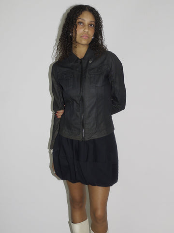 Model wearing a black fitted denim jacket with front zip and chest pockets, styled over a voluminous black mini skirt. Finished with cream-colored pointed cowboy boots, blending tough outerwear with playful western influence. Shot against a plain white wall.