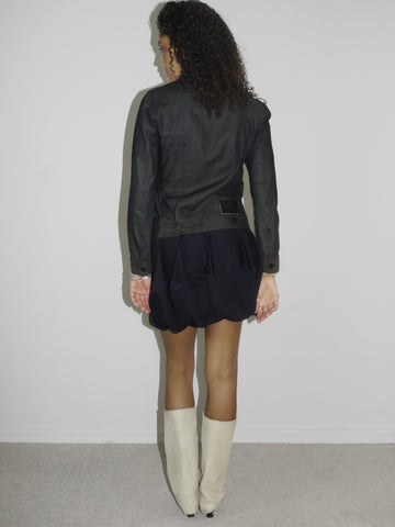 Model wearing a black fitted denim jacket with front zip and chest pockets, styled over a voluminous black mini skirt. Finished with cream-colored pointed cowboy boots, blending tough outerwear with playful western influence. Shot against a plain white wall.