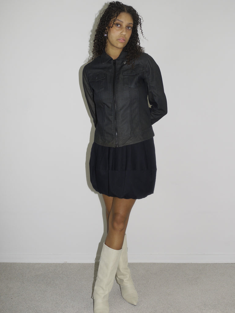 Model wearing a black fitted denim jacket with front zip and chest pockets, styled over a voluminous black mini skirt. Finished with cream-colored pointed cowboy boots, blending tough outerwear with playful western influence. Shot against a plain white wall.