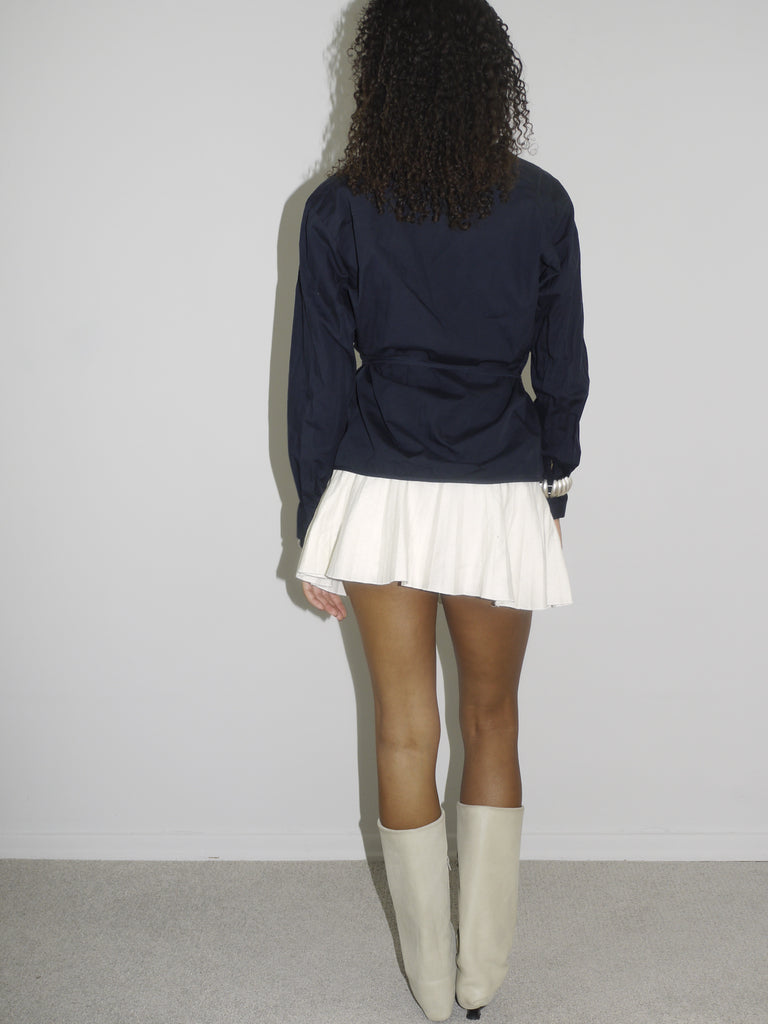 Model wearing a navy long-sleeve wrap top with a deep V-neckline, paired with a white pleated mini skirt and cream-colored pointed cowboy boots. The outfit channels a flirty, confident Y2K aesthetic with western and preppy elements. Shot against a plain white wall