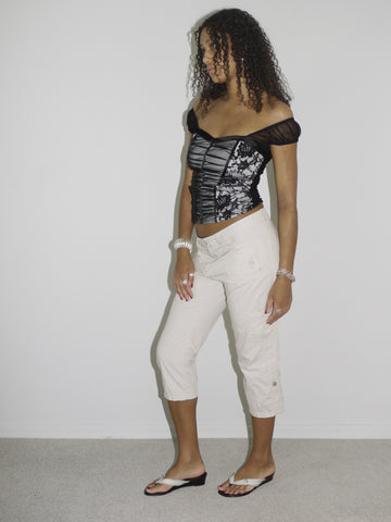 Model wearing an off-the-shoulder black satin corset top with lace-up details and floral embroidery, paired with low-rise beige capri pants. Styled with flip-flops and silver jewelry for a romantic yet edgy Y2K-inspired look. Shot against a plain white background.