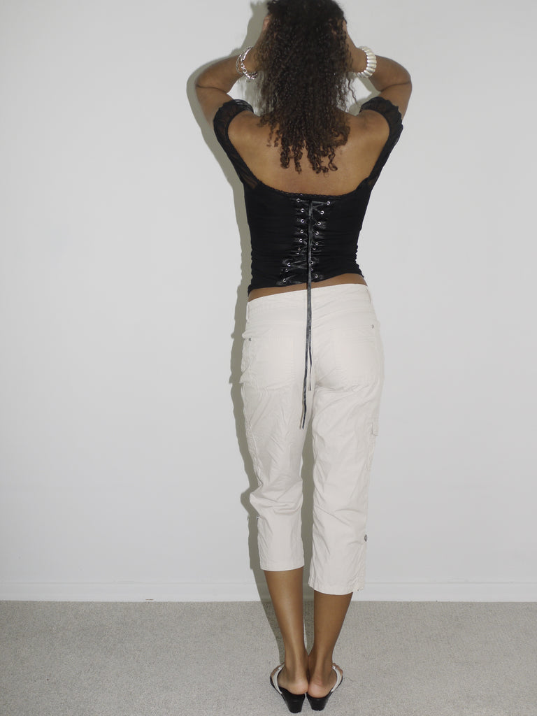 Model wearing an off-the-shoulder black satin corset top with lace-up details and floral embroidery, paired with low-rise beige capri pants. Styled with flip-flops and silver jewelry for a romantic yet edgy Y2K-inspired look. Shot against a plain white background.