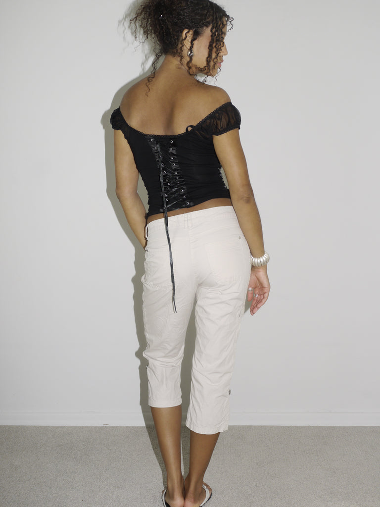 Model wearing an off-the-shoulder black satin corset top with lace-up details and floral embroidery, paired with low-rise beige capri pants. Styled with flip-flops and silver jewelry for a romantic yet edgy Y2K-inspired look. Shot against a plain white background.