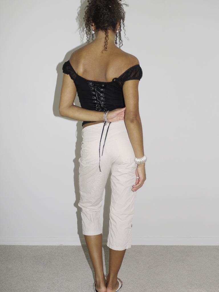 Model wearing an off-the-shoulder black satin corset top with lace-up details and floral embroidery, paired with low-rise beige capri pants. Styled with flip-flops and silver jewelry for a romantic yet edgy Y2K-inspired look. Shot against a plain white background.