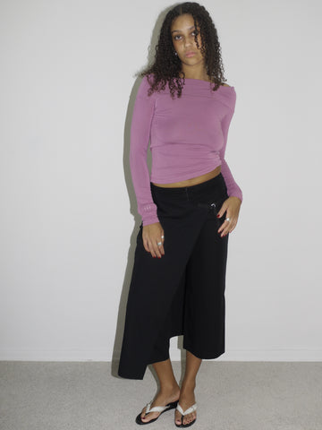 Model wearing a fitted long-sleeve top in dusty rose with a wide neckline, paired with low-rise black culotte-style trousers. Styled with flip-flops for a laid-back yet curated early-2000s look. Shot against a plain white wall.