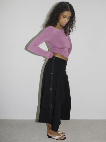 Model wearing a fitted long-sleeve top in dusty rose with a wide neckline, paired with low-rise black culotte-style trousers. Styled with flip-flops for a laid-back yet curated early-2000s look. Shot against a plain white wall.