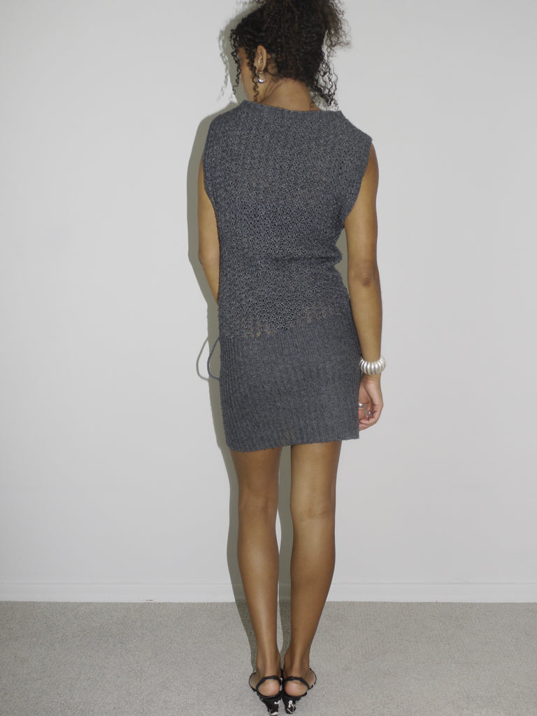 Model wearing a sleeveless grey knit mini dress with structured shoulders and subtle ribbed texture. Styled with pointed black-and-white printed heels and silver jewelry, creating a clean yet powerful vintage silhouette. Shot against a plain white background.