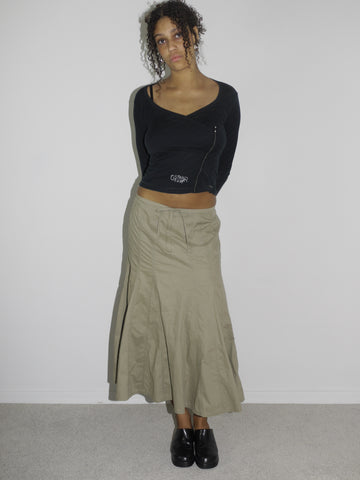 Model wearing a fitted black long-sleeve top with asymmetrical seams and subtle logo detailing, paired with a khaki-colored flared midi skirt featuring stitched paneling. Styled with chunky black slip-on shoes, evoking a utilitarian Y2K aesthetic. Shot against a plain white background.