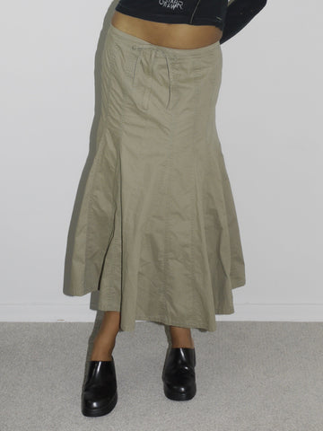 Model wearing a fitted black long-sleeve top with asymmetrical seams and subtle logo detailing, paired with a khaki-colored flared midi skirt featuring stitched paneling. Styled with chunky black slip-on shoes, evoking a utilitarian Y2K aesthetic. Shot against a plain white background.