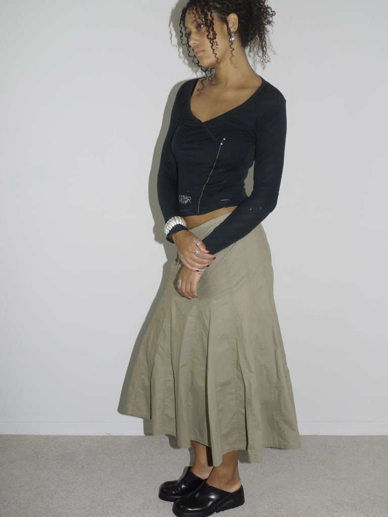 Model wearing a fitted black long-sleeve top with asymmetrical seams and subtle logo detailing, paired with a khaki-colored flared midi skirt featuring stitched paneling. Styled with chunky black slip-on shoes, evoking a utilitarian Y2K aesthetic. Shot against a plain white background.