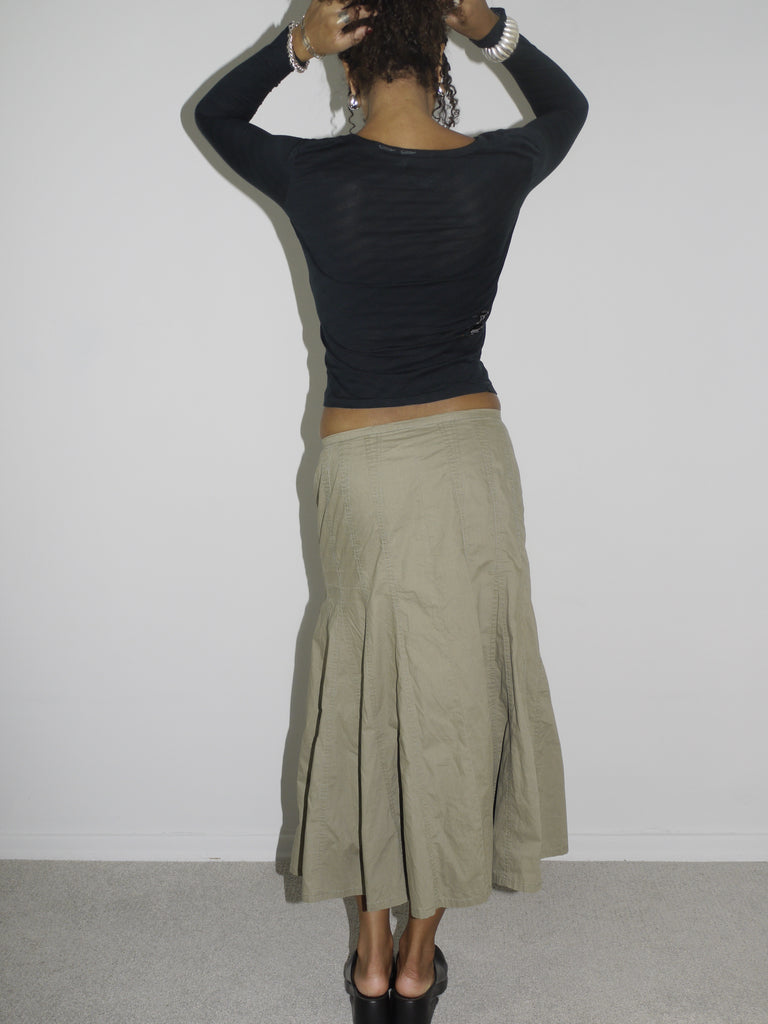 Model wearing a fitted black long-sleeve top with asymmetrical seams and subtle logo detailing, paired with a khaki-colored flared midi skirt featuring stitched paneling. Styled with chunky black slip-on shoes, evoking a utilitarian Y2K aesthetic. Shot against a plain white background.