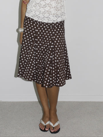 Model wearing a white sheer lace camisole with scalloped edges, paired with a brown vintage midi skirt featuring a cream polka dot pattern. Styled with flip-flops and subtle accessories, creating a soft, romantic look with early 2000s energy. Shot against a plain white wall.