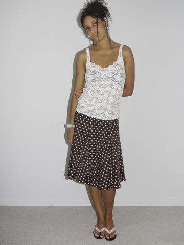 Model wearing a white sheer lace camisole with scalloped edges, paired with a brown vintage midi skirt featuring a cream polka dot pattern. Styled with flip-flops and subtle accessories, creating a soft, romantic look with early 2000s energy. Shot against a plain white wall.