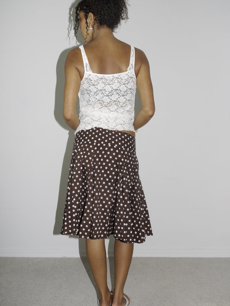 Model wearing a white sheer lace camisole with scalloped edges, paired with a brown vintage midi skirt featuring a cream polka dot pattern. Styled with flip-flops and subtle accessories, creating a soft, romantic look with early 2000s energy. Shot against a plain white wall.
