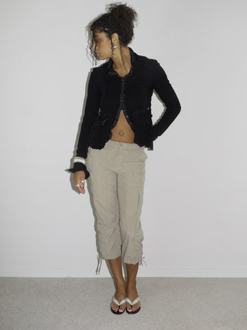Model wearing a cropped black cardigan with subtle ruffle edges and a single hook closure, paired with beige capri cargo pants featuring adjustable drawstrings at the hem. Styled with flip-flops and statement earrings, evoking a playful 2000s streetwear vibe. Shot against a plain white background.








