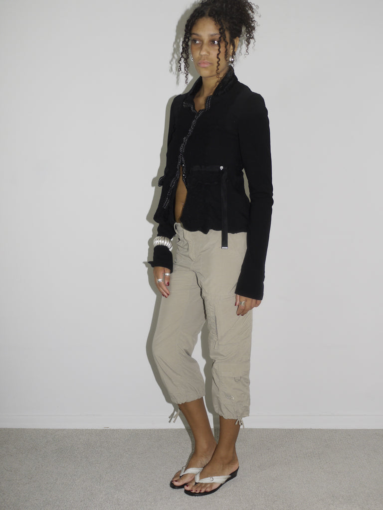 Model wearing a cropped black cardigan with subtle ruffle edges and a single hook closure, paired with beige capri cargo pants featuring adjustable drawstrings at the hem. Styled with flip-flops and statement earrings, evoking a playful 2000s streetwear vibe. Shot against a plain white background.