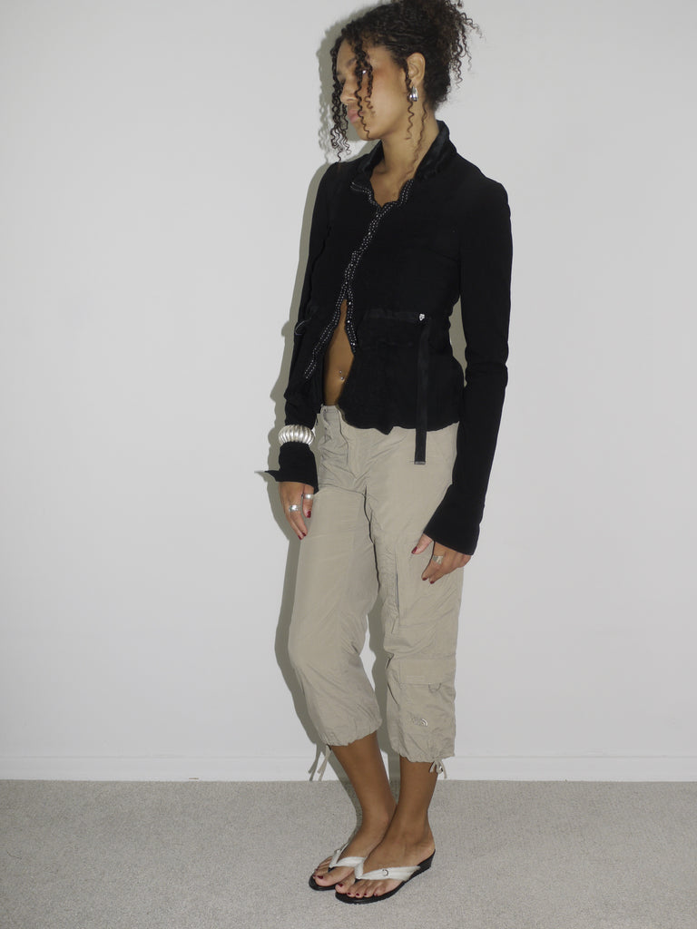 Model wearing a cropped black cardigan with subtle ruffle edges and a single hook closure, paired with beige capri cargo pants featuring adjustable drawstrings at the hem. Styled with flip-flops and statement earrings, evoking a playful 2000s streetwear vibe. Shot against a plain white background.