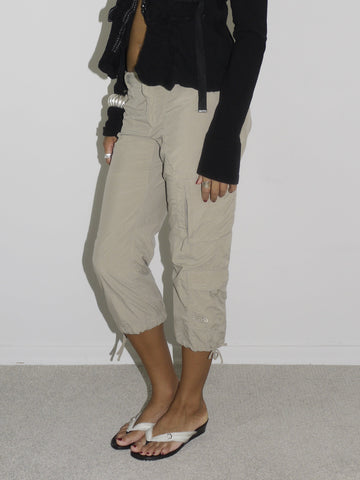 Model wearing a cropped black cardigan with subtle ruffle edges and a single hook closure, paired with beige capri cargo pants featuring adjustable drawstrings at the hem. Styled with flip-flops and statement earrings, evoking a playful 2000s streetwear vibe. Shot against a plain white background.