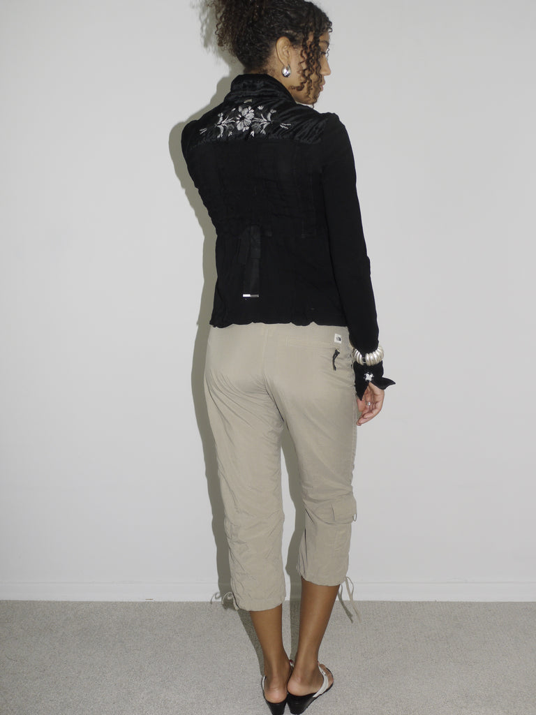 Model wearing a cropped black cardigan with subtle ruffle edges and a single hook closure, paired with beige capri cargo pants featuring adjustable drawstrings at the hem. Styled with flip-flops and statement earrings, evoking a playful 2000s streetwear vibe. Shot against a plain white background.