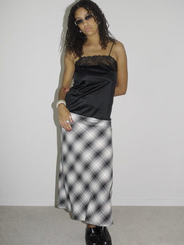 Model wearing a black satin camisole with lace trim and spaghetti straps, paired with a high-waisted black-and-white plaid midi skirt and black platform shoes. Styled with vintage Dolce & Gabbana sunglasses and silver jewelry for a bold, early-2000s-inspired look. Shot against a plain white background.