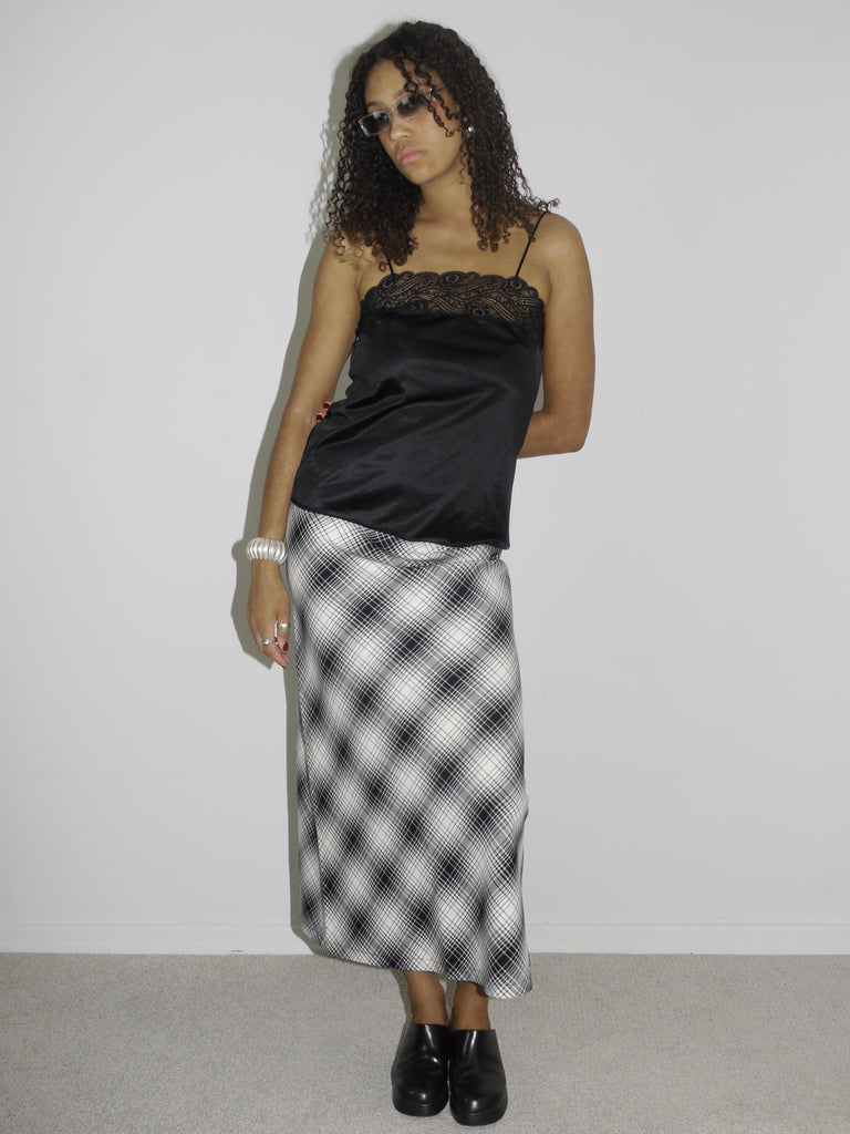 Model wearing a black satin camisole with lace trim and spaghetti straps, paired with a high-waisted black-and-white plaid midi skirt and black platform shoes. Styled with vintage Dolce & Gabbana sunglasses and silver jewelry for a bold, early-2000s-inspired look. Shot against a plain white background.