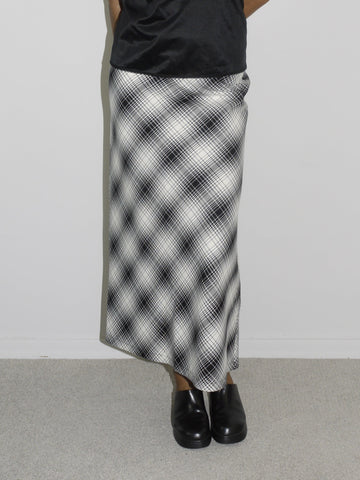 Model wearing a black satin camisole with lace trim and spaghetti straps, paired with a high-waisted, ankle-length A-line skirt in a black-and-white plaid pattern. Styled with chunky black slip-on shoes for a refined yet alternative vintage look. Shot against a white wall.