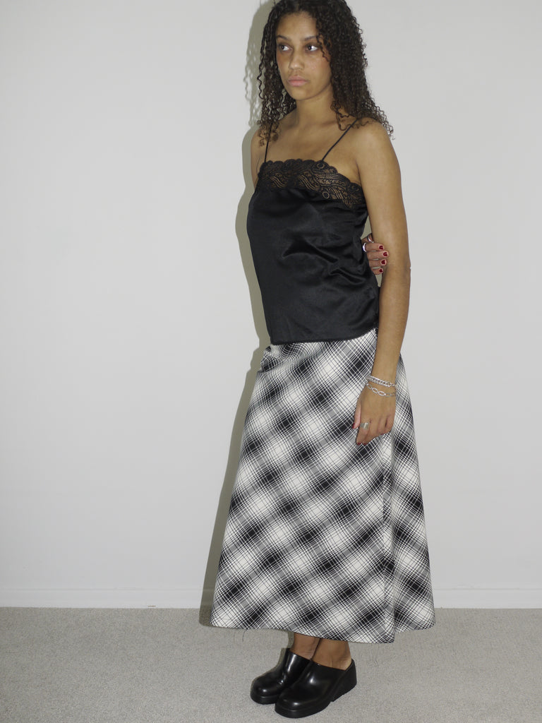 Model wearing a black satin camisole with lace trim and spaghetti straps, paired with a high-waisted, ankle-length A-line skirt in a black-and-white plaid pattern. Styled with chunky black slip-on shoes for a refined yet alternative vintage look. Shot against a white wall.