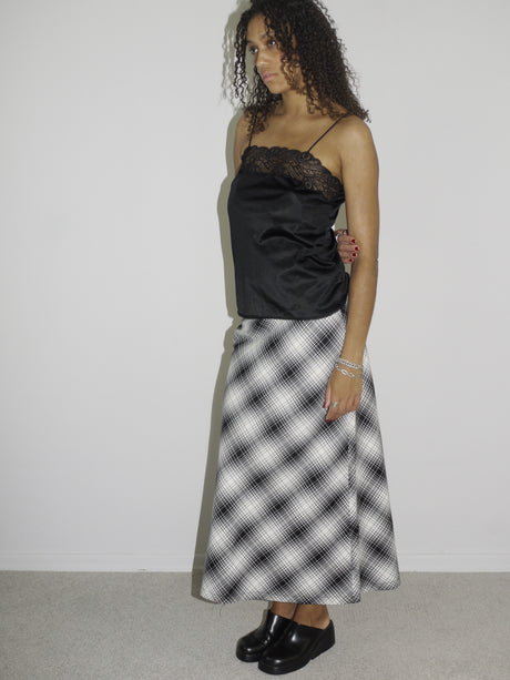 Model wearing a black satin camisole with lace trim and spaghetti straps, paired with a high-waisted, ankle-length A-line skirt in a black-and-white plaid pattern. Styled with chunky black slip-on shoes for a refined yet alternative vintage look. Shot against a white wall.