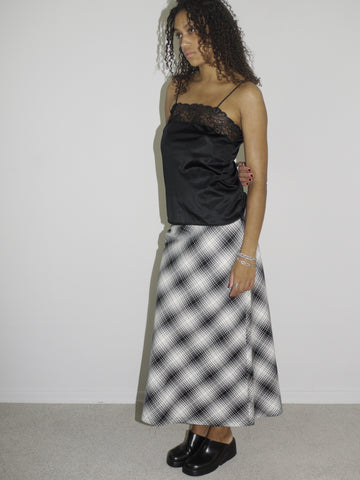Model wearing a black satin camisole with lace trim and spaghetti straps, paired with a high-waisted, ankle-length A-line skirt in a black-and-white plaid pattern. Styled with chunky black slip-on shoes for a refined yet alternative vintage look. Shot against a white wall.