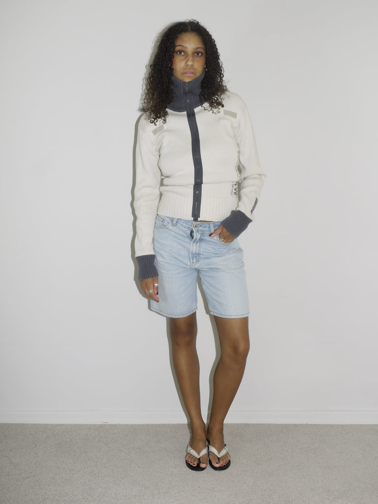 Model wearing a cream-colored zip-up ribbed sweater with black trim and lettering details on the sleeves, paired with light-wash vintage Bermuda denim shorts. Styled simply with flip-flops for a clean, early-2000s-inspired casual look. Shot against a plain white wall.