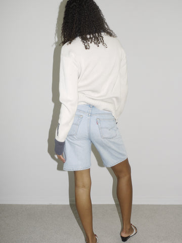 Model wearing a cream-colored zip-up ribbed sweater with black trim and lettering details on the sleeves, paired with light-wash vintage Bermuda denim shorts. Styled simply with flip-flops for a clean, early-2000s-inspired casual look. Shot against a plain white wall.