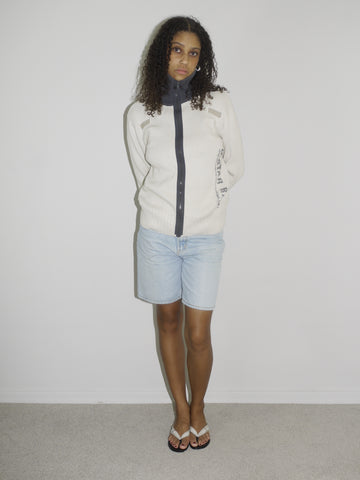 Model wearing a cream-colored zip-up ribbed sweater with black trim and lettering details on the sleeves, paired with light-wash vintage Bermuda denim shorts. Styled simply with flip-flops for a clean, early-2000s-inspired casual look. Shot against a plain white wall.