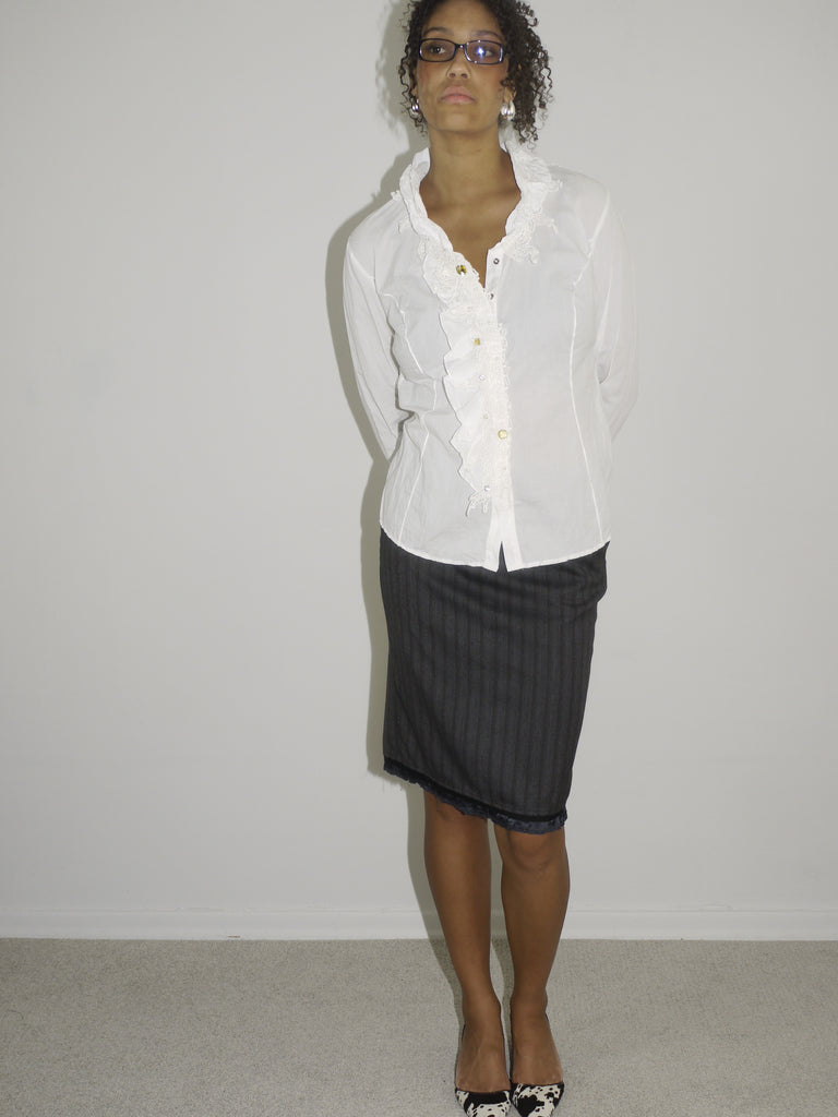 Model wearing a white vintage blouse with ruffle detailing and pearl-like buttons, paired with a grey pinstripe pencil skirt. Styled with pointed heels and slim glasses, giving off a playful take on office-chic with a retro twist. Shot against a plain white wall.