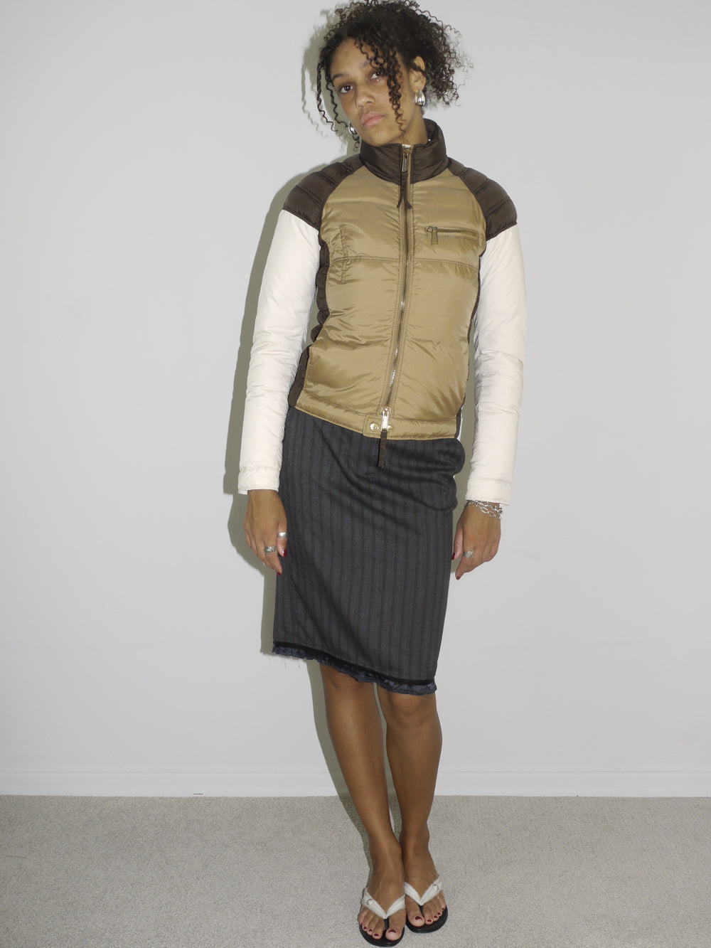 Model wearing a color-blocked puffer jacket by Dsquared² in khaki, brown, and cream tones. Styled with a pinstripe pencil skirt and flip-flops, blending sporty utility with vintage office-core energy. Shot against a white wall, hands in pockets, with an effortlessly cool posture.