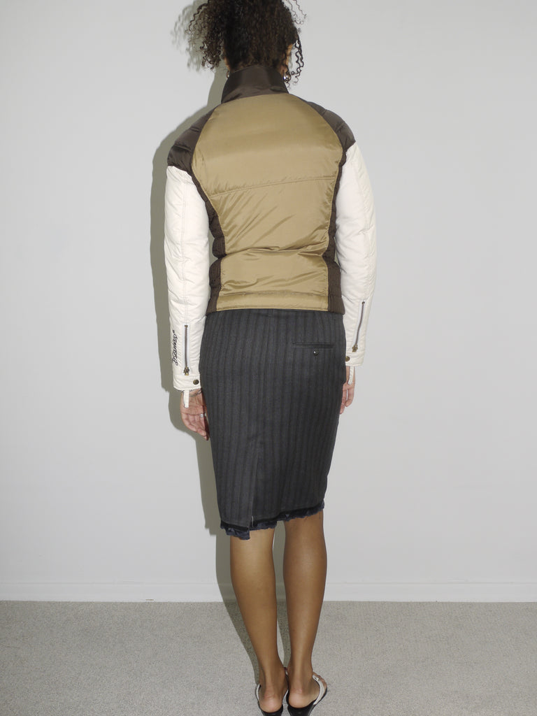 Model wearing a color-blocked puffer jacket by Dsquared² in khaki, brown, and cream tones. Styled with a pinstripe pencil skirt and flip-flops, blending sporty utility with vintage office-core energy. Shot against a white wall, hands in pockets, with an effortlessly cool posture.