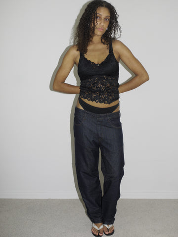 Model wearing a black lace-trimmed camisole with floral embroidery and low-rise dark wash jeans. The thong waistband is visible above the jeans, adding a bold Y2K touch. Styled with flip-flops and natural curls, shot against a plain white wall.