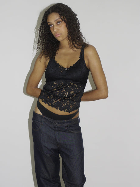 Model wearing a black lace-trimmed camisole with floral embroidery and low-rise dark wash jeans. The thong waistband is visible above the jeans, adding a bold Y2K touch. Styled with flip-flops and natural curls, shot against a plain white wall.