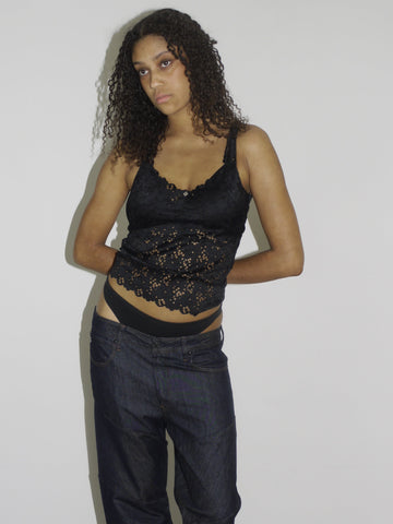 Model wearing a black lace-trimmed camisole with floral embroidery and low-rise dark wash jeans. The thong waistband is visible above the jeans, adding a bold Y2K touch. Styled with flip-flops and natural curls, shot against a plain white wall.