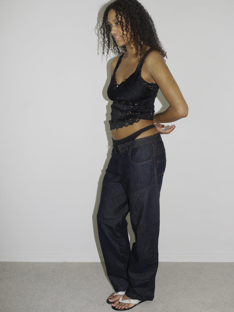 Model wearing a black lace-trimmed camisole with floral embroidery and low-rise dark wash jeans. The thong waistband is visible above the jeans, adding a bold Y2K touch. Styled with flip-flops and natural curls, shot against a plain white wall.