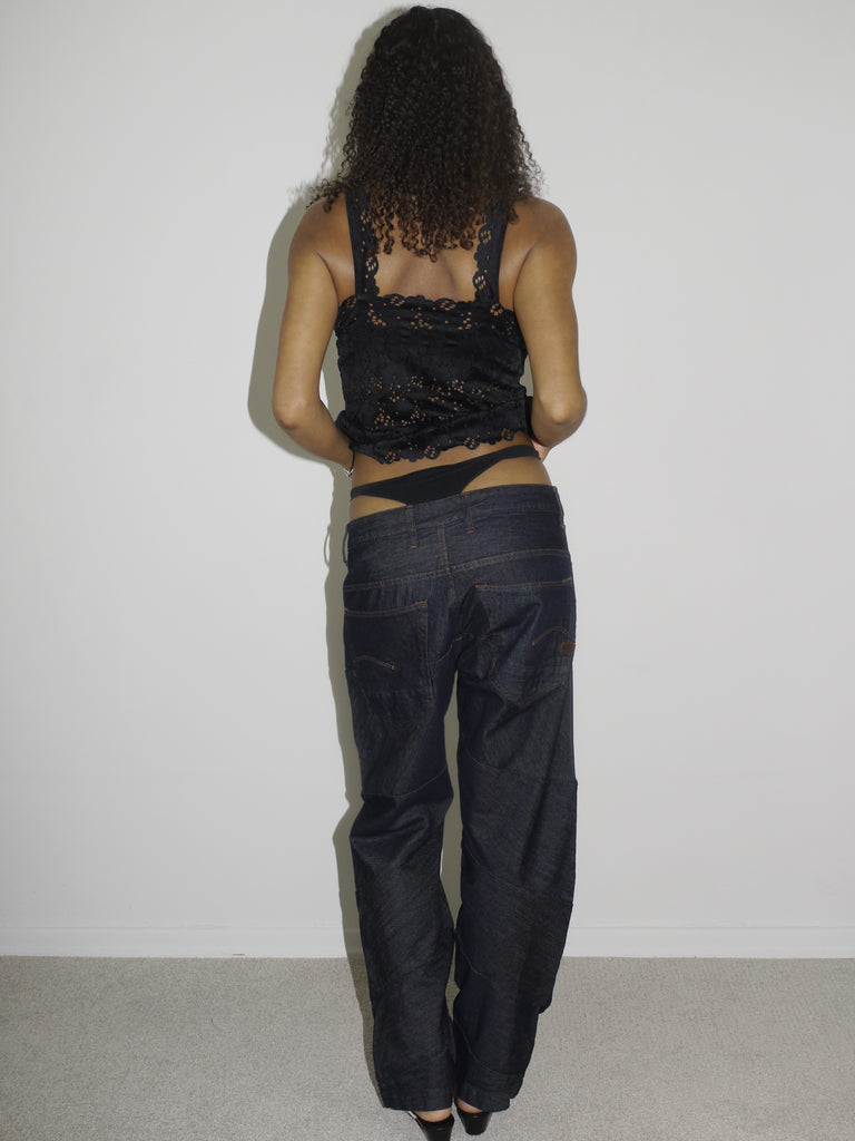 Model wearing a black lace-trimmed camisole with floral embroidery and low-rise dark wash jeans. The thong waistband is visible above the jeans, adding a bold Y2K touch. Styled with flip-flops and natural curls, shot against a plain white wall.