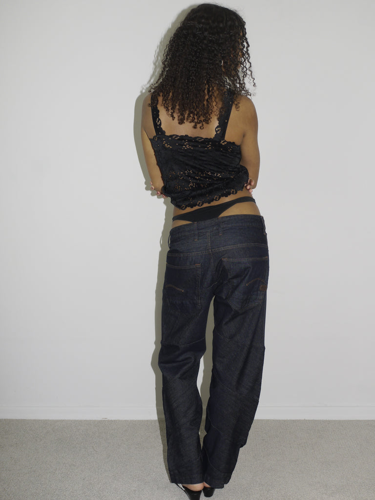 Model wearing a black lace-trimmed camisole with floral embroidery and low-rise dark wash jeans. The thong waistband is visible above the jeans, adding a bold Y2K touch. Styled with flip-flops and natural curls, shot against a plain white wall.