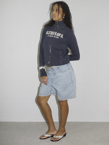 Close-up of a model wearing light blue vintage denim Bermuda shorts paired with a navy zip-up hoodie. Styled with flip-flops, silver jewelry, and a casual undone look. The outfit channels relaxed 2000s streetwear energy with a utilitarian twist.