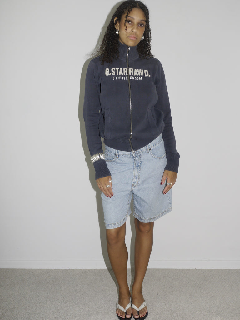 Close-up of a model wearing light blue vintage denim Bermuda shorts paired with a navy zip-up hoodie. Styled with flip-flops, silver jewelry, and a casual undone look. The outfit channels relaxed 2000s streetwear energy with a utilitarian twist.
