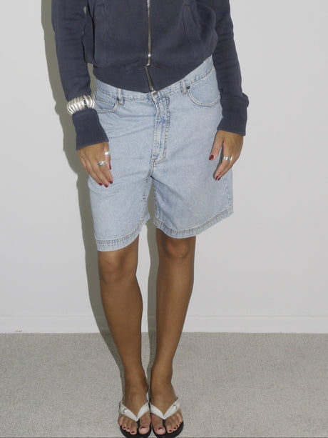 Close-up of a model wearing light blue vintage denim Bermuda shorts paired with a navy zip-up hoodie. Styled with flip-flops, silver jewelry, and a casual undone look. The outfit channels relaxed 2000s streetwear energy with a utilitarian twist.