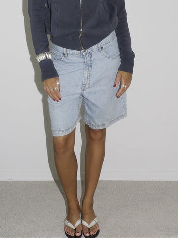 Close-up of a model wearing light blue vintage denim Bermuda shorts paired with a navy zip-up hoodie. Styled with flip-flops, silver jewelry, and a casual undone look. The outfit channels relaxed 2000s streetwear energy with a utilitarian twist.