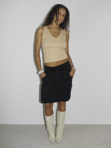 Model wearing a beige ribbed tank top and a low-rise black skirt with drawstring detail, styled with pointed cream-colored cowboy boots. The look is early 2000s-inspired, blending casual minimalism with a touch of western edge. Shot against a plain white background.