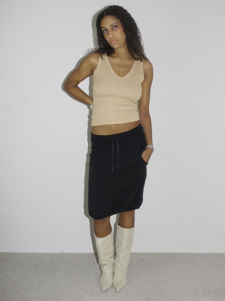 Model wearing a beige ribbed tank top and a low-rise black skirt with drawstring detail, styled with pointed cream-colored cowboy boots. The look is early 2000s-inspired, blending casual minimalism with a touch of western edge. Shot against a plain white background.